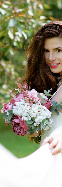 A woman in a white dress holding a bouquet of pink and white flowers, surrounded by greenery and smiling at the camera.