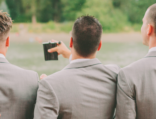 Three men in suits stand with their backs to the camera, one holding a flask, in an outdoor setting with greenery.