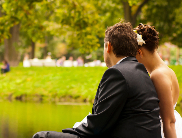 A couple, dressed formally, sits by a pond surrounded by greenery, enjoying a peaceful moment outdoors.